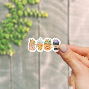 May include: Four Halloween-themed coffee drinks with different toppings. The drinks are in clear cups with lids and straws. The toppings include whipped cream, sprinkles, and a pumpkin face.