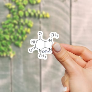 Puede incluir: Una pegatina blanca con un dibujo de línea negra de la estructura química de la cafeína. El texto "Caffeine" está escrito en azul debajo de la estructura.