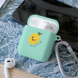 Könnte beinhalten: Eine hellgrüne AirPod-Hülle mit einem niedlichen Cartoon-Zitronendesign. Die Zitrone hat ein zwinkerndes Gesicht und der Text "Squeeze the Day!" ist darunter gedruckt. Die Hülle hat einen silbernen Karabinerhaken.