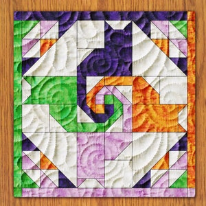 May include: A colorful quilt block with a geometric design. The block is made up of squares and triangles in shades of green, purple, orange, and white. The fabric has a quilted texture.