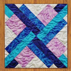 Puede incluir: Un bloque de colcha con un patrón geométrico de tela azul, rosa y blanca. La tela tiene un efecto tie-dye y está acolchada con una puntada decorativa.