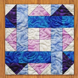 May include: A quilt block with a geometric pattern in shades of blue, pink, and purple. The block is made up of squares and triangles, and the fabric has a swirl pattern.