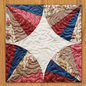 May include: A quilt block featuring a star design in the centre. The star is surrounded by eight triangles in alternating colours of red, blue, and brown. The triangles are made of fabric with floral patterns.