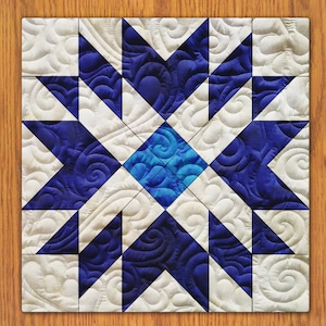 Op de afbeelding: Een quiltblok met een stervormig ontwerp in blauw en wit. De ster is gemaakt van kleinere driehoeken en vierkanten. De achtergrond is wit met een draaiend patroon.