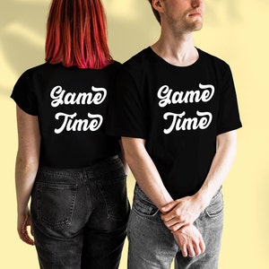 May include: Two people wearing black t-shirts with the text "Game Time" in white. The person on the left is wearing a black t-shirt with the text on the back. The person on the right is wearing a black t-shirt with the text on the front.