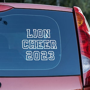 May include: A red car with a white vinyl sticker on the back window that says "Lion Cheer 2023".