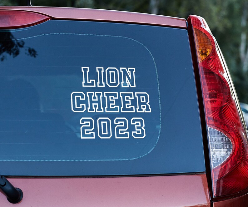 May include: A red car with a white vinyl sticker on the back window that says "Lion Cheer 2023".