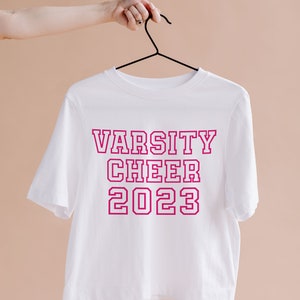 May include: White short-sleeved t-shirt with the text "VARSITY CHEER 2023" in pink outlined letters.