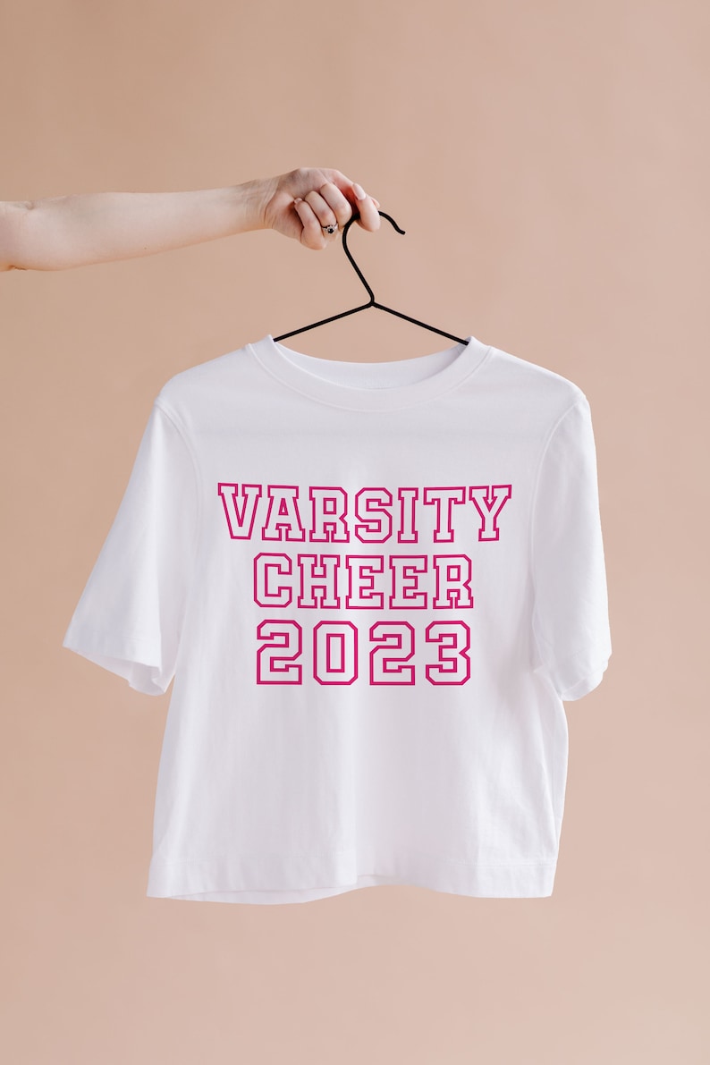 May include: White short-sleeved t-shirt with the text "VARSITY CHEER 2023" in pink outlined letters.