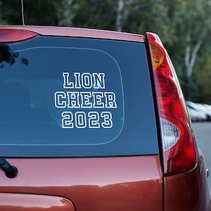 May include: A red car with a white vinyl sticker on the back window that says "Lion Cheer 2023".