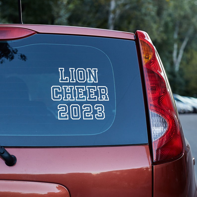 May include: A red car with a white vinyl sticker on the back window that says "Lion Cheer 2023".