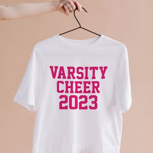 May include: White t-shirt with pink lettering that reads "Varsity Cheer 2023" hanging on a black hanger.
