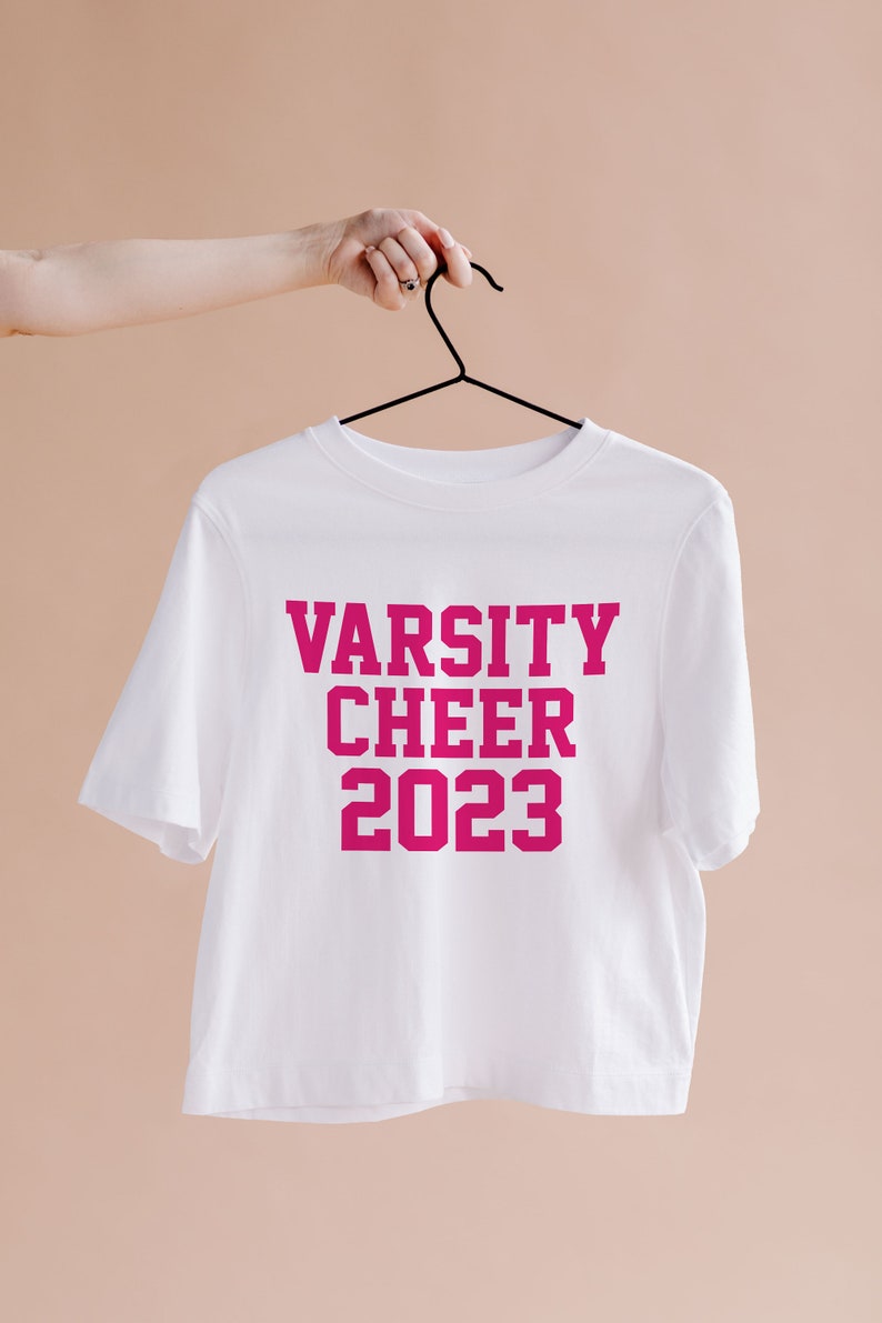 May include: White t-shirt with pink lettering that reads "Varsity Cheer 2023" hanging on a black hanger.