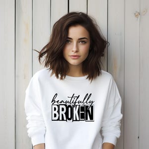 Pu&ograve; includere: Felpa bianca con la scritta "beautifully broken" stampata in nero. Il testo &egrave; in grassetto, in un carattere a blocchi.