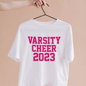 May include: White cropped t-shirt with the text "VARSITY CHEER 2023" in pink lettering.