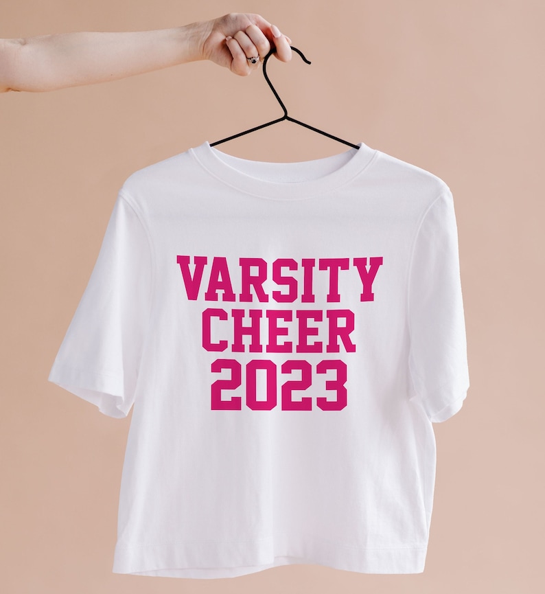 May include: White cropped t-shirt with the text "VARSITY CHEER 2023" in pink lettering.