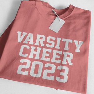 May include: A pink t-shirt with the text "VARSITY CHEER 2023" in white.