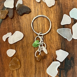 May include: A silver keyring with three sea glass charms in green, brown, and white. The keyring is surrounded by various pieces of sea glass in shades of white, brown, and blue-green, arranged on a wooden surface.