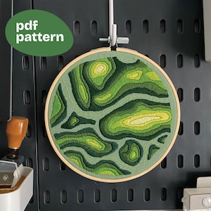 May include: A round embroidery hoop featuring a layered, topographical design in shades of green and yellow. The text "pdf pattern" is in a green oval. The artwork is mounted on a dark gray pegboard.
