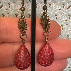 Peut inclure: Boucles d'oreilles en forme de goutte rouge avec des motifs sculptés complexes. Les boucles d'oreilles sont rehaussées d'un charme de fleur en filigrane doré.