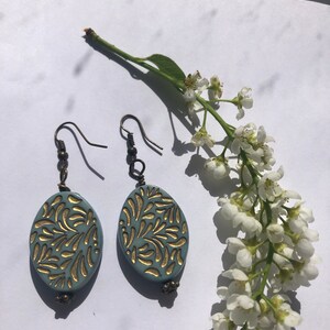 Puede incluir: Un par de pendientes ovalados azules y dorados con un diseño floral. Los pendientes cuelgan de un gancho plateado.