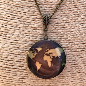 Puede incluir: Un collar de cadena de bronce con un colgante que presenta un diseño de mapa del mundo. El colgante tiene forma ovalada y tiene un fondo marrón oscuro con un mapa del mundo de color dorado.