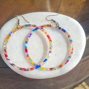 Puede incluir: Un par de pendientes de aro coloridos con un patrón de remolino multicolor. Los pendientes están hechos de un material plástico transparente y tienen un gancho de color cobre.