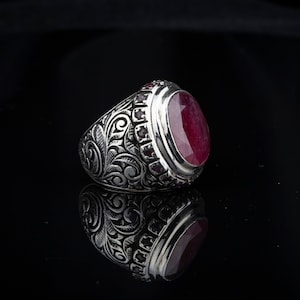 Può includere: Un anello d'argento con una gemma ovale rosso intenso. L'anello presenta intricate incisioni floreali e a volute sulla fascia e intorno alla gemma. Piccole gemme rosse sono incastonate attorno alla pietra principale.