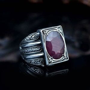 May include: A silver ring featuring an intricate design and a large, faceted, ruby-red gemstone set in a square bezel.