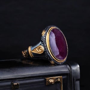Può includere: Un anello d'argento con una grande pietra ovale di colore rosso scuro incastonata in una lunetta dorata. L'anello presenta disegni intricati e dettagliati sulla fascia.