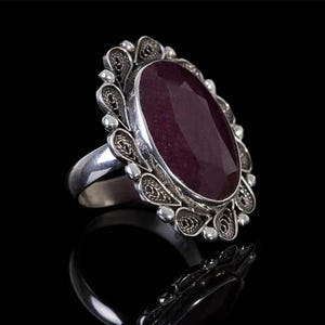 May include: A silver ring with an oval-shaped, dark purple gemstone set in a filigree design.
