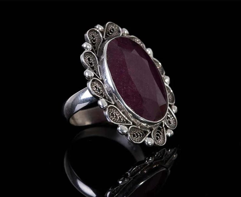 May include: A silver ring with an oval-shaped, dark purple gemstone set in a filigree design.