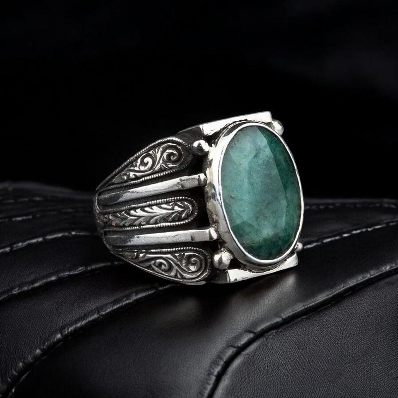 May include: A silver ring with an oval-shaped green gemstone set in a detailed silver band. The band features intricate scrollwork and a textured design.