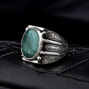 May include: A silver ring with an oval-shaped green gemstone set in a detailed silver band. The band features intricate scrollwork and floral designs.