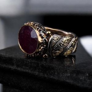 Könnte beinhalten: Ein silberner Ring mit einem großen, runden, dunkelroten Edelstein, der in einer goldenen Fassung sitzt. Der Ringarm ist mit filigranen, gravierten Blumenmustern verziert.