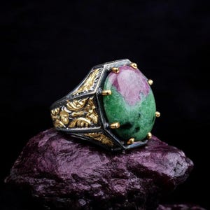 May include: A silver ring with a green and pink gemstone set in a gold-toned ornate setting. The ring is resting on a dark purple rock.