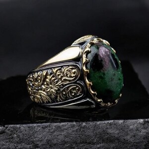 Può includere: Un anello d'argento con una pietra preziosa verde e rosa incastonata in una montatura d'oro. L'anello presenta un intricato design floreale.