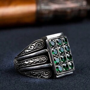 May include: A silver ring with an intricate design and a rectangular green gemstone setting. The gemstone is set with multiple smaller green stones.
