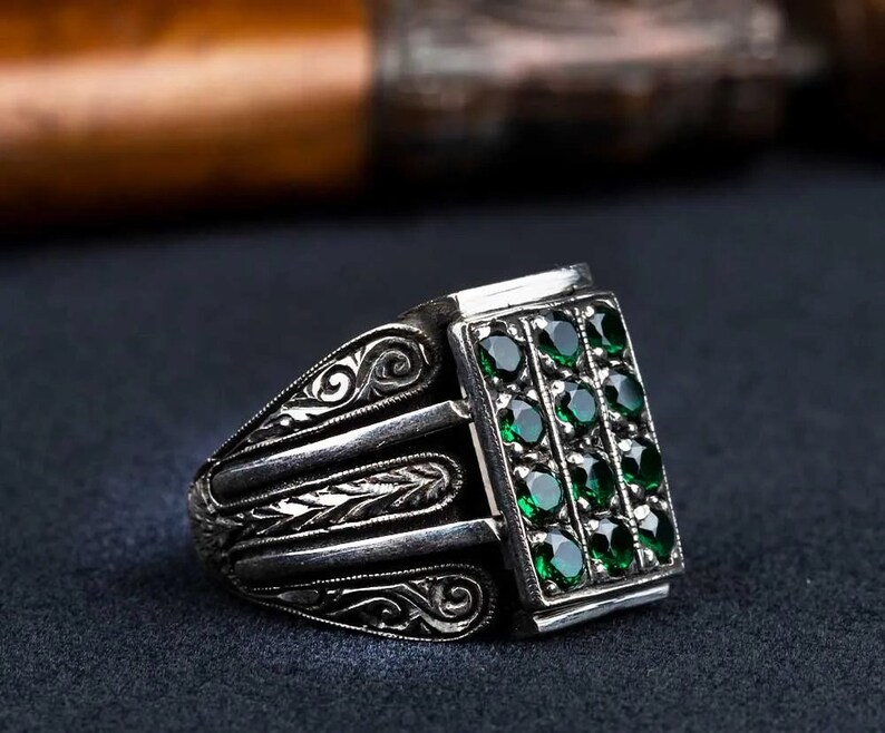 May include: A silver ring with an intricate design and a rectangular green gemstone setting. The gemstone is set with multiple smaller green stones.