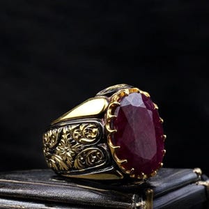 May include: A gold-toned ring with an oval, deep red gemstone. The ring features intricate floral and scrollwork designs on the band. The gemstone is the focal point, set in a detailed bezel.