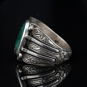 May include: A silver ring with an oval-shaped green gemstone set in a bezel setting. The band is intricately engraved with floral designs.