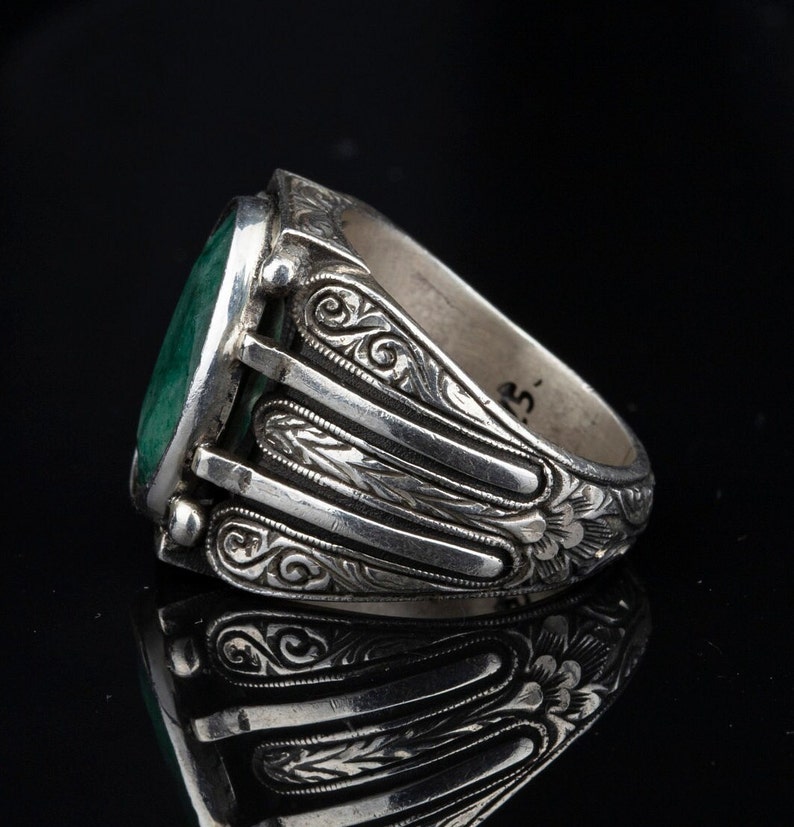 May include: A silver ring with an oval-shaped green gemstone set in a bezel setting. The band is intricately engraved with floral designs.