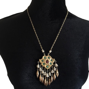 Puede incluir: Un collar con colgante dorado con un diseño floral. El colgante presenta piedras preciosas turquesas y rojas, con dijes colgantes en forma de lágrima. El collar se exhibe en un maniquí negro.