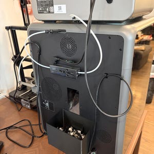May include: The back view of a gray 3D printer featuring a black panel with two circular ventilation grilles, a rectangular waste bin attached at the bottom containing plastic scraps, and various black and white cables routed across the surface.