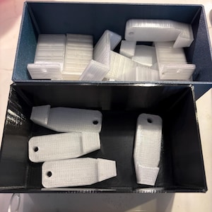 May include: Two rectangular storage bins, one dark blue and one black, filled with multiple white 3D-printed plastic components. Each component features a flat rectangular base, a small circular hole at one end, and a protruding hook-like tab.