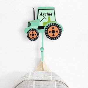 May include: A light green tractor-shaped wall hook with the name "Archie" printed on it. The hook is holding a gray and white plaid backpack. The tractor has orange and black wheels and a green body.