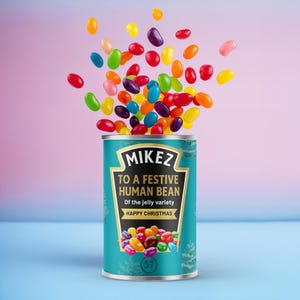 Puede incluir: Una lata turquesa de gominolas "Mikez" con el texto "To a Festive Human Bean" y "Happy Christmas". Se muestran gominolas de colores saliendo de la lata sobre un fondo rosa y azul.