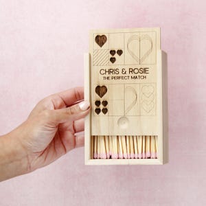 Può includere: Scatola di fiammiferi in legno con un cassetto scorrevole. La parte anteriore della scatola presenta un motivo a forma di cuore e il testo "Chris & Rosie The Perfect Match". Il cassetto è aperto e pieno di fiammiferi.