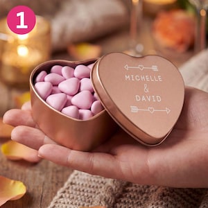 May include: A rose gold heart-shaped tin filled with pink heart-shaped sweets. The lid is open, revealing the sweets, and is engraved with the names "MICHELLE & DAVID" and arrow and heart designs. The tin is held in a person's hand.