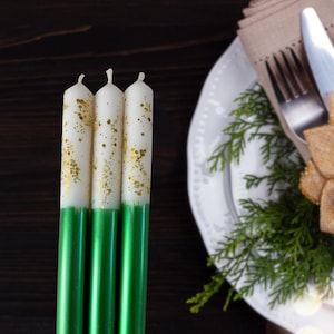 Op de afbeelding: Drie witte kaarsen met gouden glitter en groene voetjes. De kaarsen staan op een donker houten oppervlak.